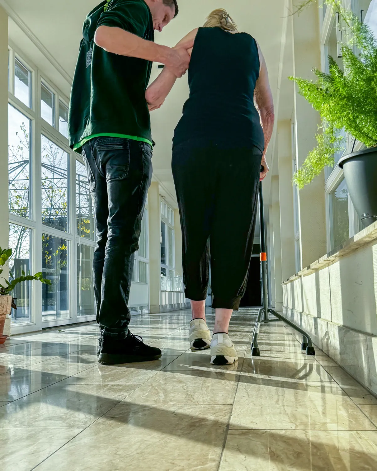 Flur einer Pflegeeinrichtung, ein Therapeut begleitet eine Bewohnerin mit Rollator, von hinten gesehen